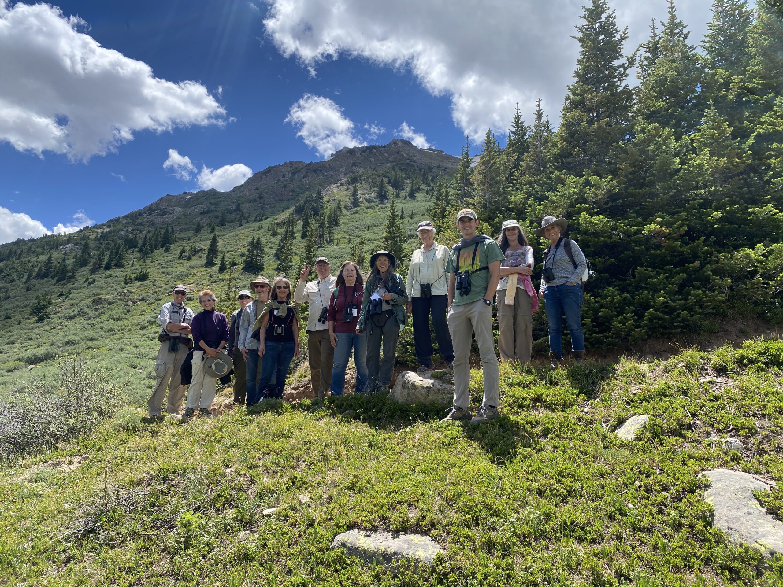 Group photo of 12 birders on a green high alpine slope. All have binoculars around their necks.