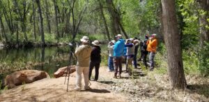 Group photo of 11 birders looking out over still water in a plains riparian habitat. Many birders have their binoculars raised to their eyes.